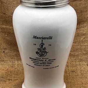 May include: A white ceramic wine bottle with a silver rim and lid. The bottle has black text that reads "Masciarelli" and "Montepulciano d, Abrozzo". The bottle is 750 ml.