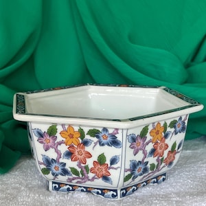 May include: A white octagonal ceramic planter with a floral design in shades of blue, orange, pink, and green. The planter has a scalloped edge and a blue and white wave pattern around the bottom.
