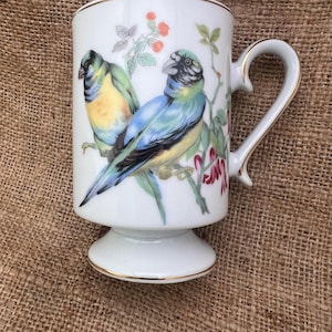 May include: White ceramic pedestal mug with a gold rim and handle. The mug features a colorful illustration of two birds perched on a flowering branch. The birds have blue, yellow, and green plumage. The handle is curved and the base is round.