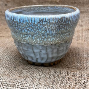 May include: A handmade ceramic bowl with a textured exterior and a light blue glaze that drips down the sides. The bowl has a wide opening and a rounded shape, perfect for serving or display.