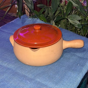 May include: A terracotta casserole dish with a wooden handle and a lid. The dish is a light brown color and the lid is a darker brown color.