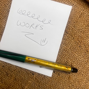May include: A green and gold pen with the text "Runnin' in the Sun ARIZONA" is next to a white notepad with the word "WORKS" written on it. The pen is lying on a brown burlap surface.