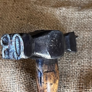 May include: Close-up of a vintage hammer with a dark, textured metal head and a worn wooden handle. The hammer head has a stamped design. The handle shows signs of age and use, with a natural wood grain.