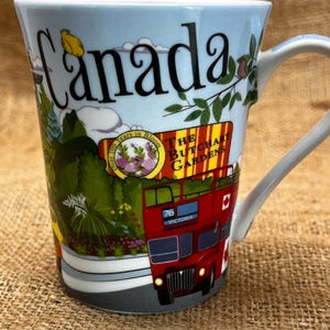 May include: A ceramic mug with a light blue sky background and the word "Canada" in black. The mug features a red double-decker bus, trees, and the text "The Butchart Gardens".