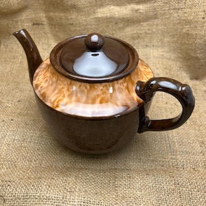 May include: A brown ceramic teapot with a matching lid and handle. The teapot has a rounded body with a lighter brown, textured pattern on the upper portion. The spout and handle are a darker shade of brown.