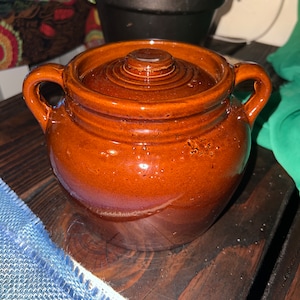 May include: A brown ceramic crock pot with a lid. The crock pot has two handles on the sides and a slightly rounded shape.