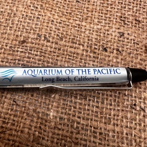 May include: A silver pen with a black tip and a silver clip. The pen has the text "AQUARIUM OF THE PACIFIC Long Beach, California" printed on it. The pen is lying on a brown, textured surface.