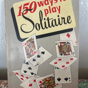 May include: A vintage book titled "150 Ways to Play Solitaire" with a gray cover and playing cards scattered around the title. The cards are in various suits and numbers, including a Jack of Hearts, a Queen of Diamonds, and a 10 of Clubs.