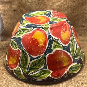 May include: A ceramic bowl with a wide, flared shape. The bowl is decorated with a pattern of red apples with yellow centers and green leaves against a dark teal background. The rim is brown.