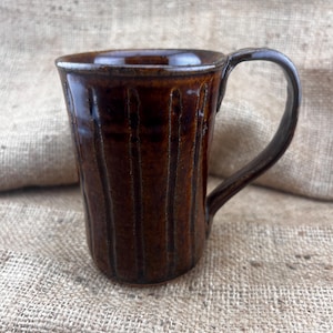 May include: A brown ceramic mug with vertical lines and a handle. The mug is sitting on a burlap surface.