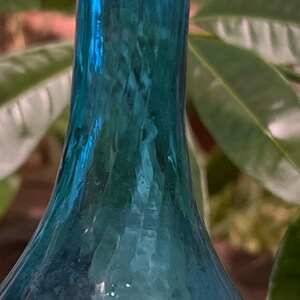 Puede incluir: Una botella de vidrio azul verdoso con fondo redondeado y cuello largo. La botella tiene una superficie texturizada y parece ser antigua.