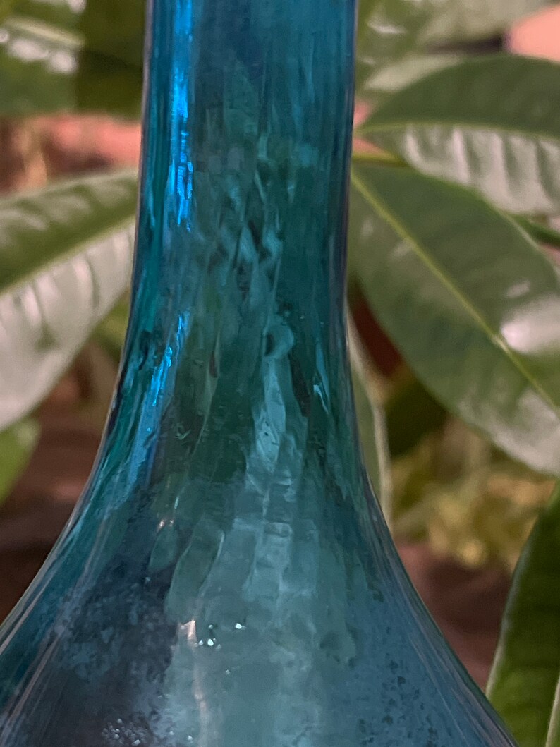 Puede incluir: Una botella de vidrio azul verdoso con fondo redondeado y cuello largo. La botella tiene una superficie texturizada y parece ser antigua.