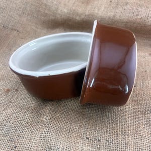 May include: Two small, oval-shaped brown ceramic bowls with a glossy finish. The interior of each bowl is white. The bowls are resting on a textured, tan fabric background. The bowls are likely for serving individual portions of food.