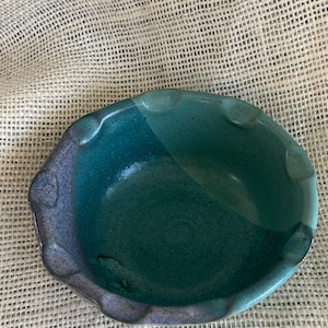 Puede incluir: Un pequeño cuenco de cerámica azul y verde con un borde festoneado. El cuenco está esmaltado y tiene un acabado moteado.