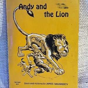 May include: A yellow book cover with the title "Andy and the Lion" in black text. The cover features a black and white illustration of a lion with a boy and a dog. The text "Story and pictures by JAMES DAUGHERTY" is at the bottom of the cover.