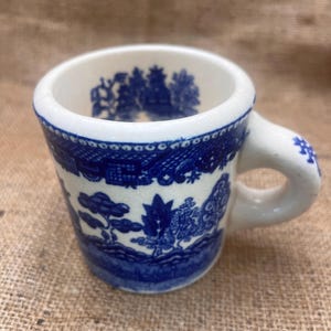 May include: A small, white ceramic mug with a blue Willow pattern design. The mug features a rounded handle and a blue band around the rim. The blue pattern depicts a landscape scene. The mug is approximately 2.5 inches tall.