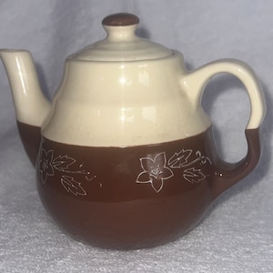 May include: A small brown and cream ceramic teapot with a floral design. The teapot has a lid and a handle.
