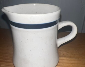 Puede incluir: Crema de cerámica blanca con una banda azul alrededor de la parte superior y moteada en todas partes. La crema tiene un asa en el lado derecho.