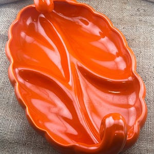 May include: Orange ceramic serving dish with three divided sections and two handles. The dish has a glossy finish and a leaf-like design.