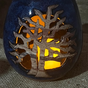May include: A blue ceramic candle holder with a tree design cut out. The tree is silhouetted against a warm orange glow from the candle inside. The candle holder has a textured surface.