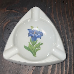 May include: A white triangular ceramic ashtray with a blue and green flower design.