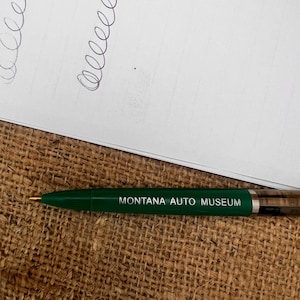 May include: A green ballpoint pen with the text "MONTANA AUTO MUSEUM" printed in white. The pen is lying on a burlap surface next to a piece of paper with black cursive writing.