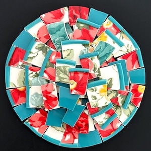 May include: A circular mosaic arrangement composed of numerous ceramic shards. The pieces feature a floral pattern with red, yellow, and green tones on a white background, interspersed with solid teal-colored ceramic fragments.