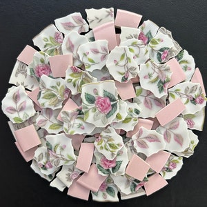 May include: A circular arrangement of broken porcelain pieces and pink tiles. The porcelain shards feature a floral pattern with pink roses, green leaves, and gold trim. The pink tiles are rectangular and scattered among the porcelain.