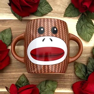 May include: A brown ceramic mug featuring a sock monkey face design with a white muzzle, large red lips, and black eyes. The mug has a textured surface resembling knit fabric and includes two side handles, one on each side of the monkey face.