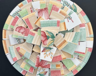 May include: A circular mosaic coaster composed of broken ceramic tiles. The tiles feature floral and geometric patterns in shades of beige, red, green, and yellow. The coaster is set against a black background.