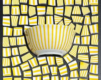 May include: A ceramic bowl featuring a repeating pattern of vertical yellow and white stripes. The bowl is centered against a background composed of numerous small, square-shaped ceramic tiles, each displaying a variety of yellow and white striped designs.