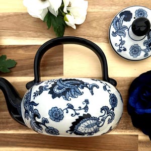May include: A white and blue floral patterned teapot with a black handle and spout. The lid, also white with a blue floral design, sits beside the teapot. The teapot is on a wooden surface with white flowers.