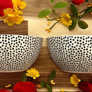 May include: Two white ceramic bowls with a black polka dot pattern. The bowls are surrounded by red and yellow flowers and greenery, set against a wooden background. The bowls are ideal for serving food.
