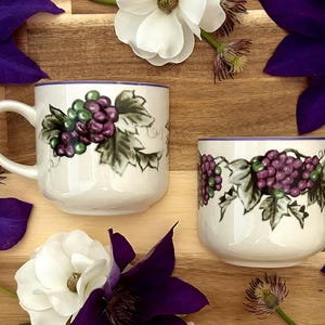 May include: Two white ceramic mugs with a grape design. One mug has a handle, and the other is a tumbler. The grape design features green and purple grapes with green leaves. The mugs are surrounded by purple and white flowers.