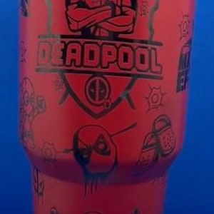 May include: A red tumbler featuring a black Deadpool design with the text "WHERE IS FRANCIS?" and "DEADPOOL". The tumbler has a black lid and is displayed on a blue stand. The bottom of the tumbler has the text "Syn's Creations".