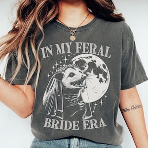 May include: A gray t-shirt with a graphic of a possum wearing a veil and a flower crown. The text on the shirt reads "In My Feral Bride Era".