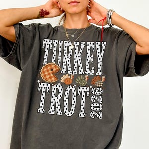 May include: A charcoal gray short-sleeve t-shirt featuring the text "TURKEY TROT 2026" in white letters with black polka dots. The graphic includes a brown plaid pie slice and three stylized turkeys with orange, green, and brown patterns.