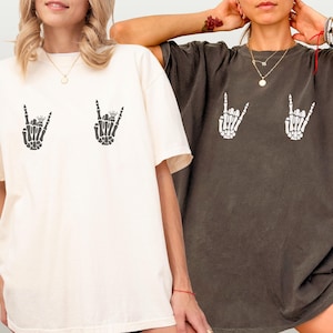 May include: Two oversized t-shirts are shown, one ivory and one pepper-coloured. Each shirt has a graphic of a skeleton hand making a rock-and-roll gesture. The text "IVORY" and "PEPPER" are at the bottom of the image.