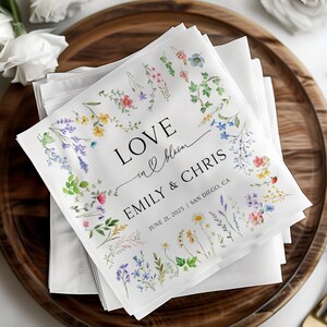 May include: White wedding napkins with a floral design and the text "LOVE in bloom EMILY & CHRIS JUNE 21, 2025 | SAN DIEGO, CA"