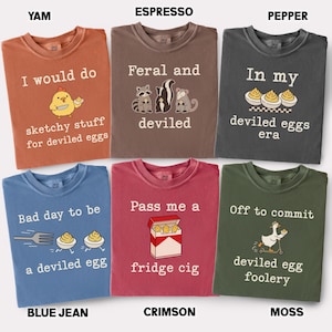 May include: Six crewneck t-shirts in yam, espresso, pepper, blue jean, crimson, and moss colors. Each shirt features white text and graphic illustrations related to deviled eggs, including a chicken, raccoons, a skunk, an opossum, and a goose.