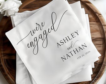 May include: White paper napkins with black text that reads "we're engaged" in a cursive font. Below the text are the names "Ashley and Nathan" and the date "Feb 26, 2026".