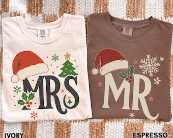 Può includere: Due magliette, una avorio e una espresso, con disegni natalizi. La maglietta avorio mostra "MRS" con cappello di Babbo Natale, albero e fiocchi di neve. La maglietta espresso mostra "MR" con cappello di Babbo Natale e fiocco di neve. Le magliette riportano "IVORY" e "ESPRESSO".