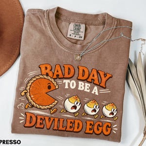 May include: An espresso-colored short-sleeve t-shirt featuring a graphic of a pumpkin pie slice chasing three deviled eggs. The text reads "BAD DAY TO BE A DEVILED EGG" in orange letters. The shirt has a Comfort Colors brand label in the neck area.