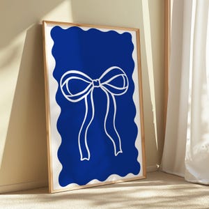 May include: A framed art print featuring a white bow design on a vibrant blue background. The bow is centered, with flowing ribbons. The blue background has a wavy, irregular border. The frame is a light wood color.