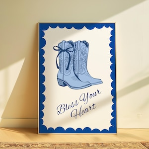 May include: A framed print featuring light blue cowboy boots with decorative stitching and a bow. The boots are set against a cream background with the phrase "Bless Your Heart" in a script font. The frame is a dark blue scalloped edge.