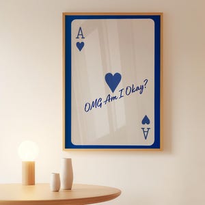 May include: Framed wall art featuring a playing card design. The card is white with blue accents, including the Ace symbol and the text "OMG Am I Okay?" The frame is a light wood color with a blue border.