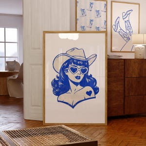 May include: A blue and white pop art style print of a woman wearing a cowboy hat and heart-shaped sunglasses. The print is in a wooden frame and is displayed with other framed prints featuring cowboy boots. The artwork is displayed in a room with wooden floors.
