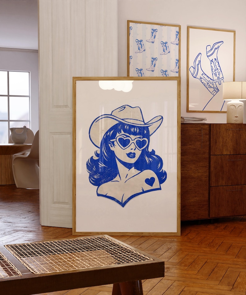 May include: A blue and white pop art style print of a woman wearing a cowboy hat and heart-shaped sunglasses. The print is in a wooden frame and is displayed with other framed prints featuring cowboy boots. The artwork is displayed in a room with wooden floors.