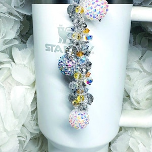 May include: A decorative charm for a tumbler, featuring a silver chain with iridescent crystal beads and three sparkling, round, crystal-covered accents. The charm is attached to a white tumbler with the brand name "STANLEY" visible.