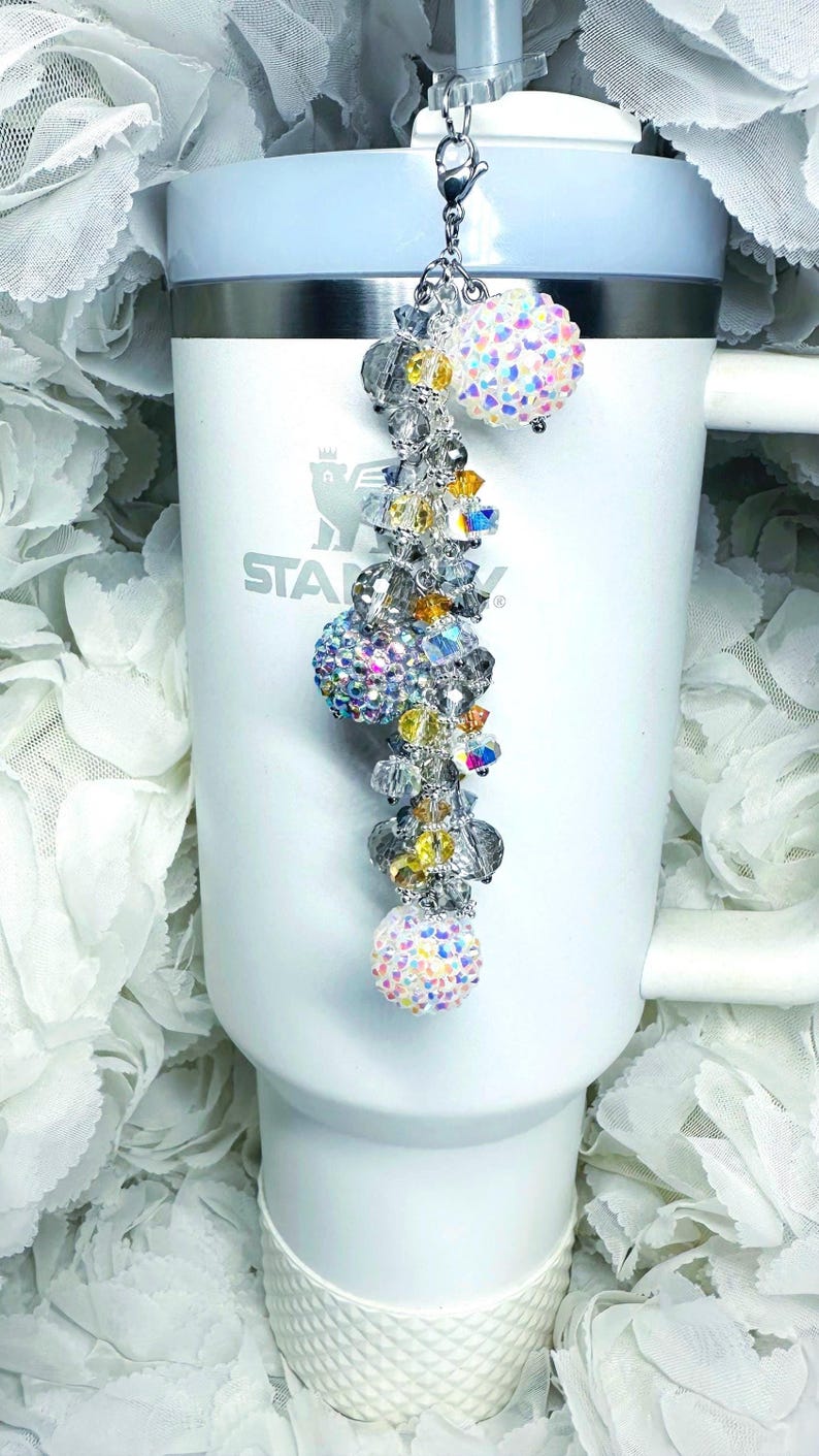 May include: A decorative charm for a tumbler, featuring a silver chain with iridescent crystal beads and three sparkling, round, crystal-covered accents. The charm is attached to a white tumbler with the brand name "STANLEY" visible.